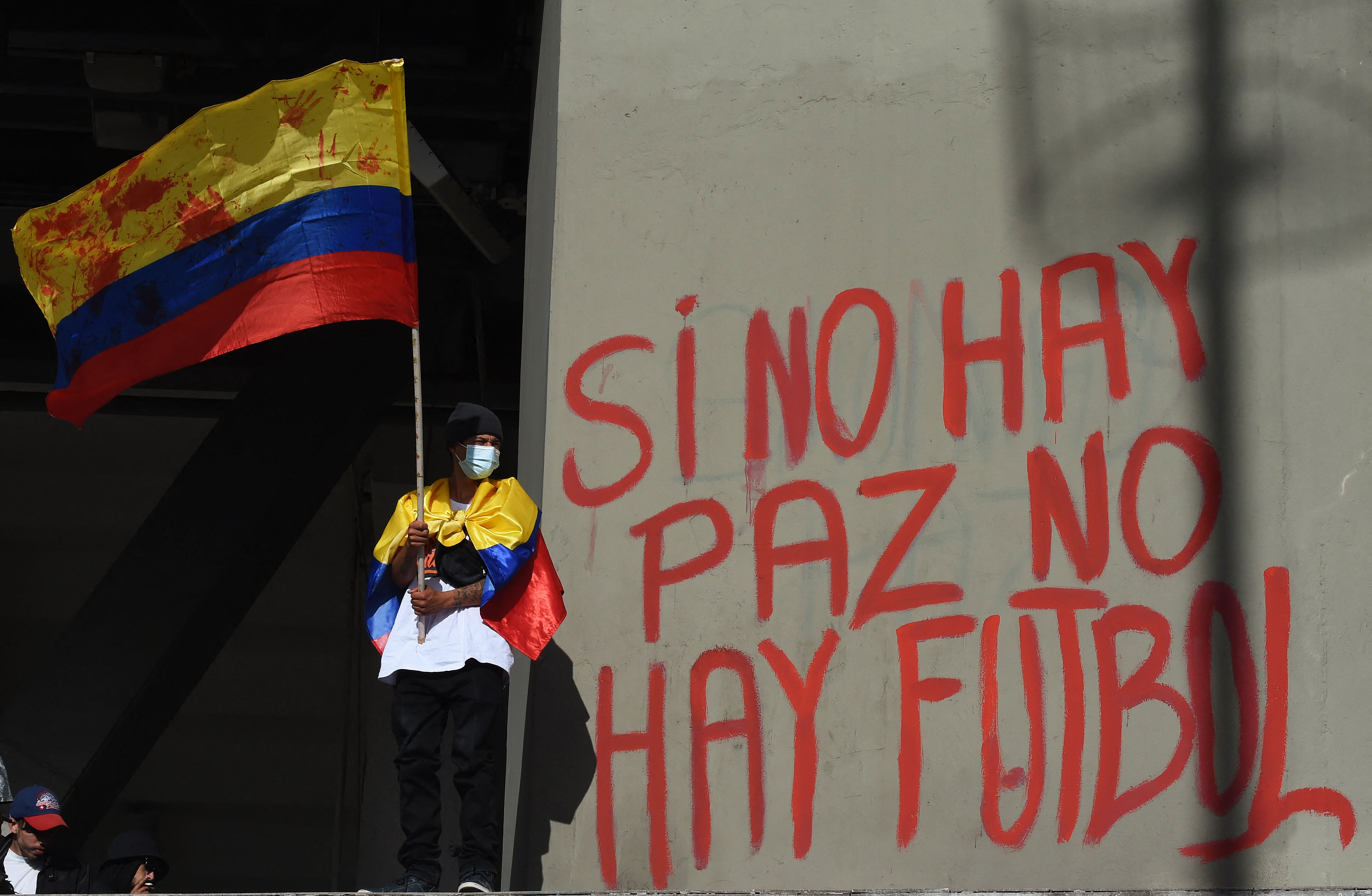 Un manifestante sostiene una bandera nacional colombiana junto a un graffiti en una pared del estadio El Campín que dice 'No hay paz, no hay fútbol' durante una protesta contra Colombia como sede del torneo de fútbol Copa América frente al estadio El Campín en Bogotá, el 19 de mayo de 2021 mientras el país enfrenta su peor crisis política y civil en la historia reciente.